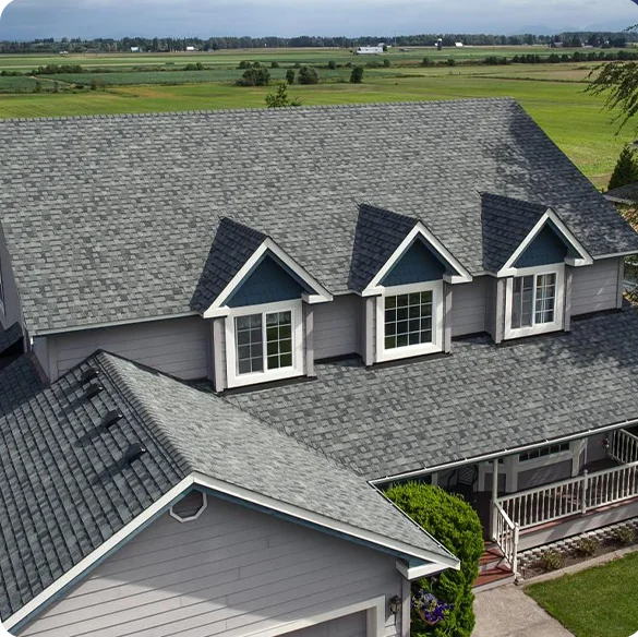 Aerial view of a large grey house with an asphalt shingle roof and white dormer windows in a green countryside location.